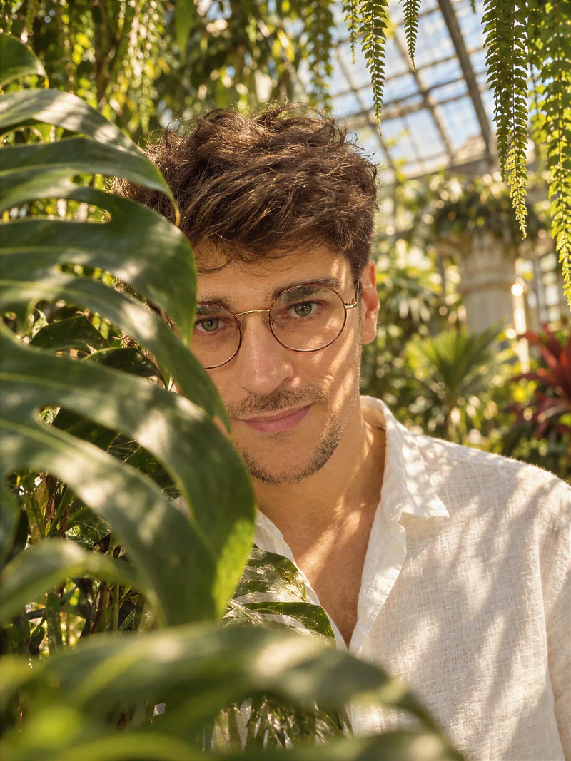 Andrey in round glasses and a white linen shirt, framed by monstera leaves in a sunlit greenhouse